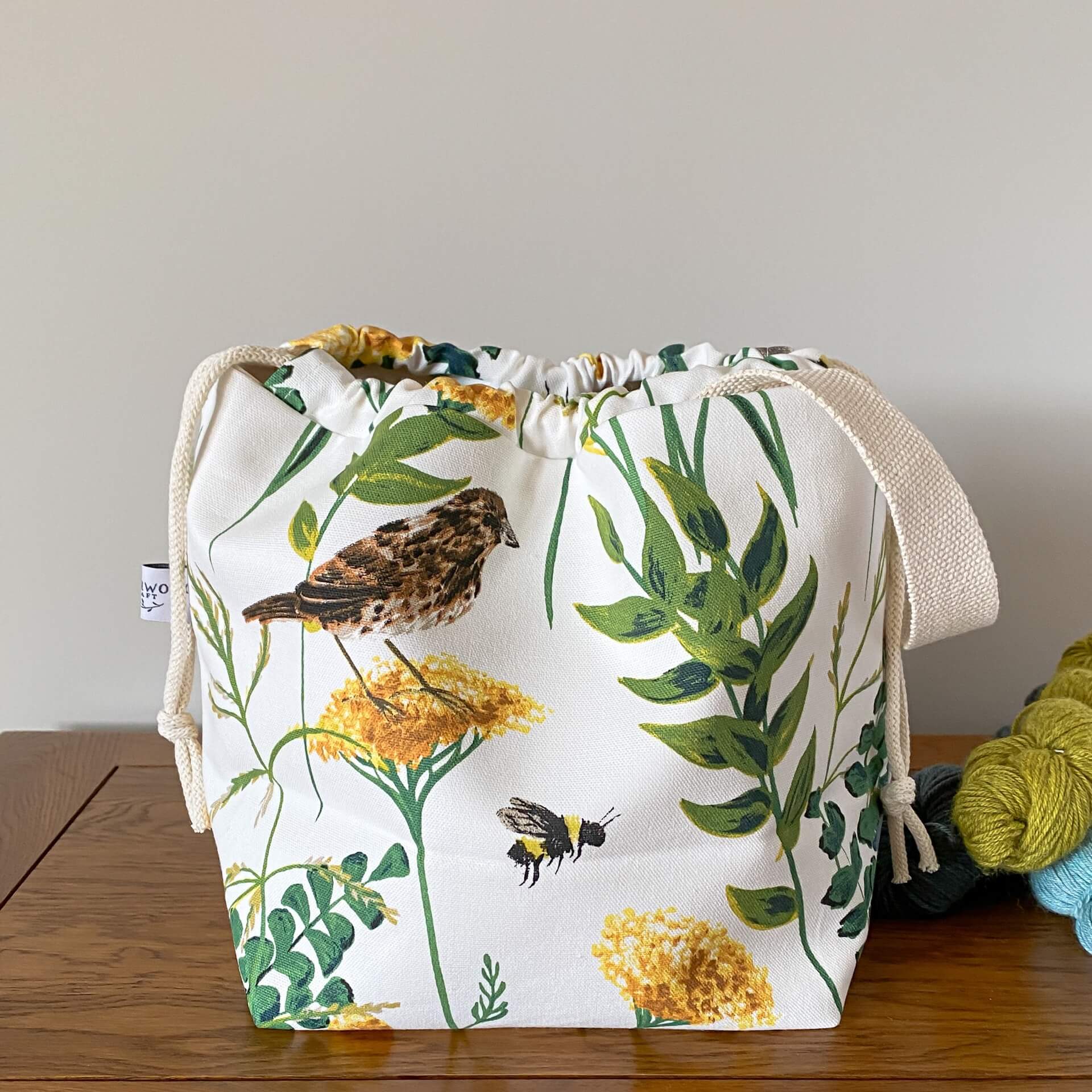 A drawstring bag used for organising knitting and crochet projects sits on a wooden table. Next to it are three skeins of yarn in shades of blue and green. The bag has been made using a white fabric that has a summery print with foliage, a song thrush bird and a bumble bee printed on it. 