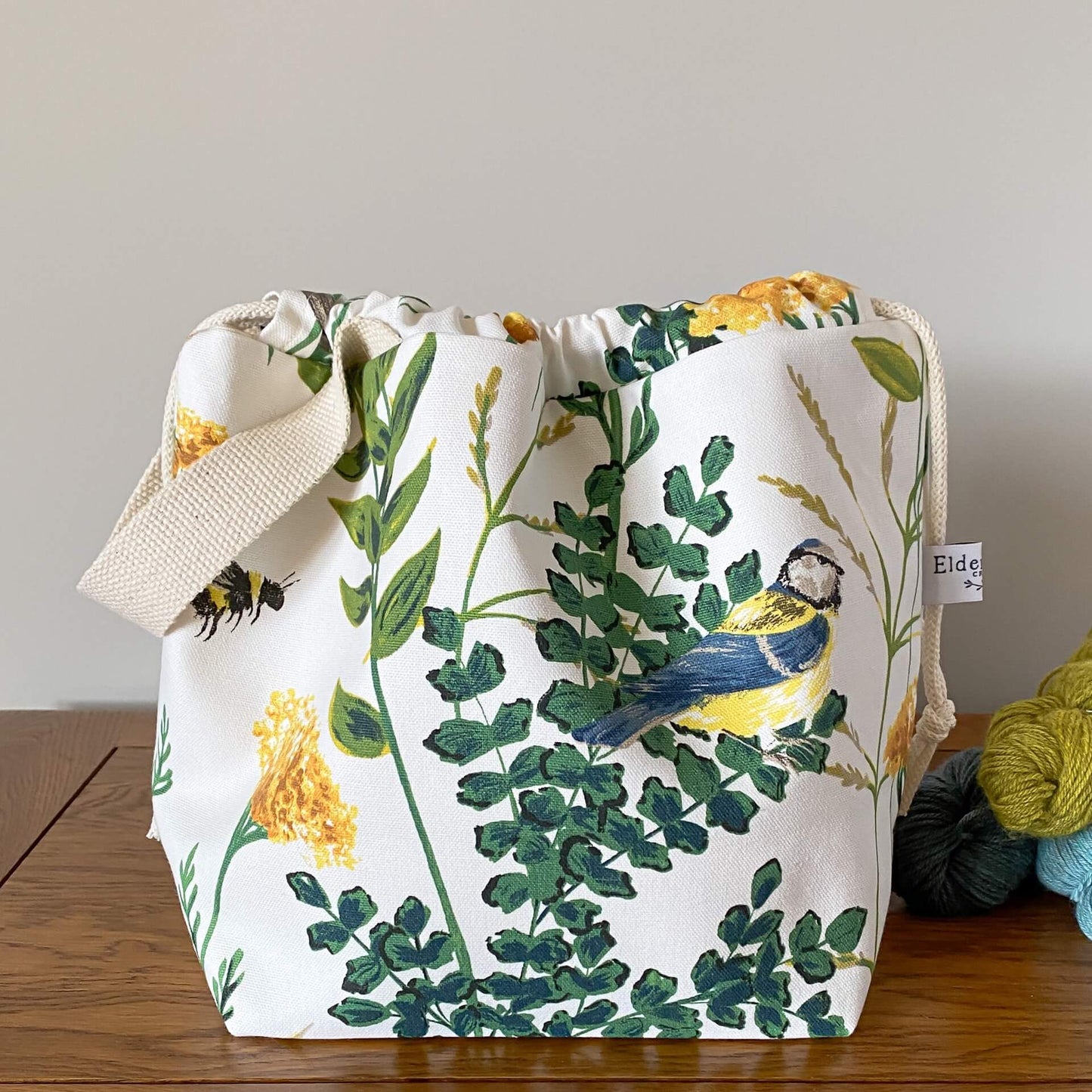 A drawstring bag used for organising knitting and crochet projects sits on a wooden table. Next to it are three skeins of yarn in shades of blue and green. The bag has been made using a white fabric that has a summery print with foliage, a blue tit bird and a bumble bee printed on it.