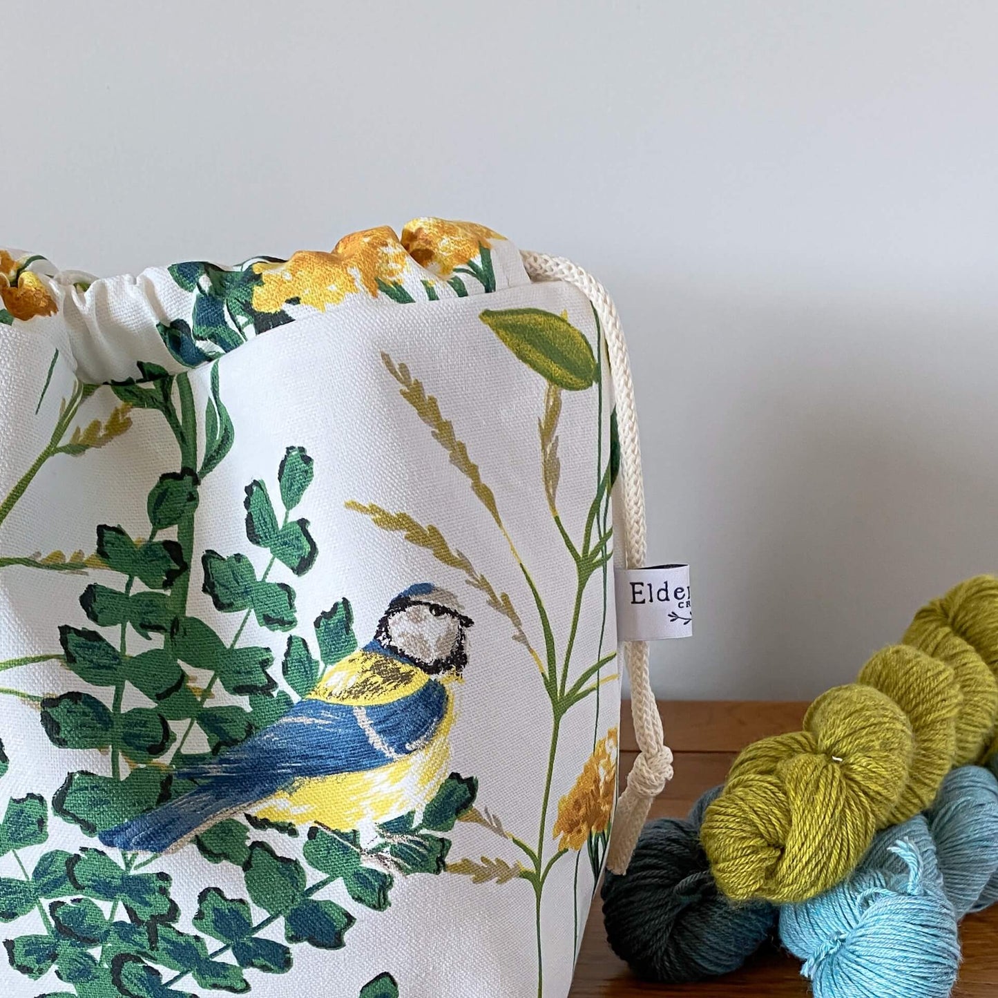 A close up of part of a knitting project bag showing a blue tit bird. Also seen on a table behind the bag are some skeins of yarn.