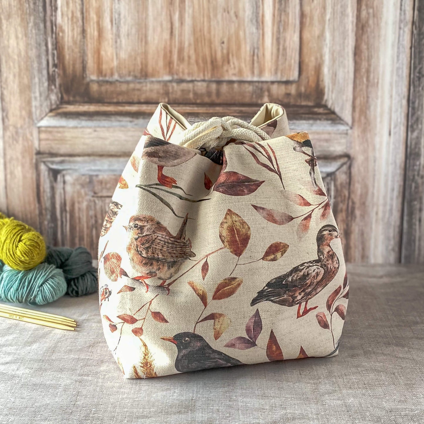 A fabric knitting project bag with a print of birds and autumn leaves in warm, earthy tones. The bag is cinched at the top with a drawstring, and behind it, there is a partially visible wooden background. A few skeins of yarn and knitting needles are placed nearby on the surface.