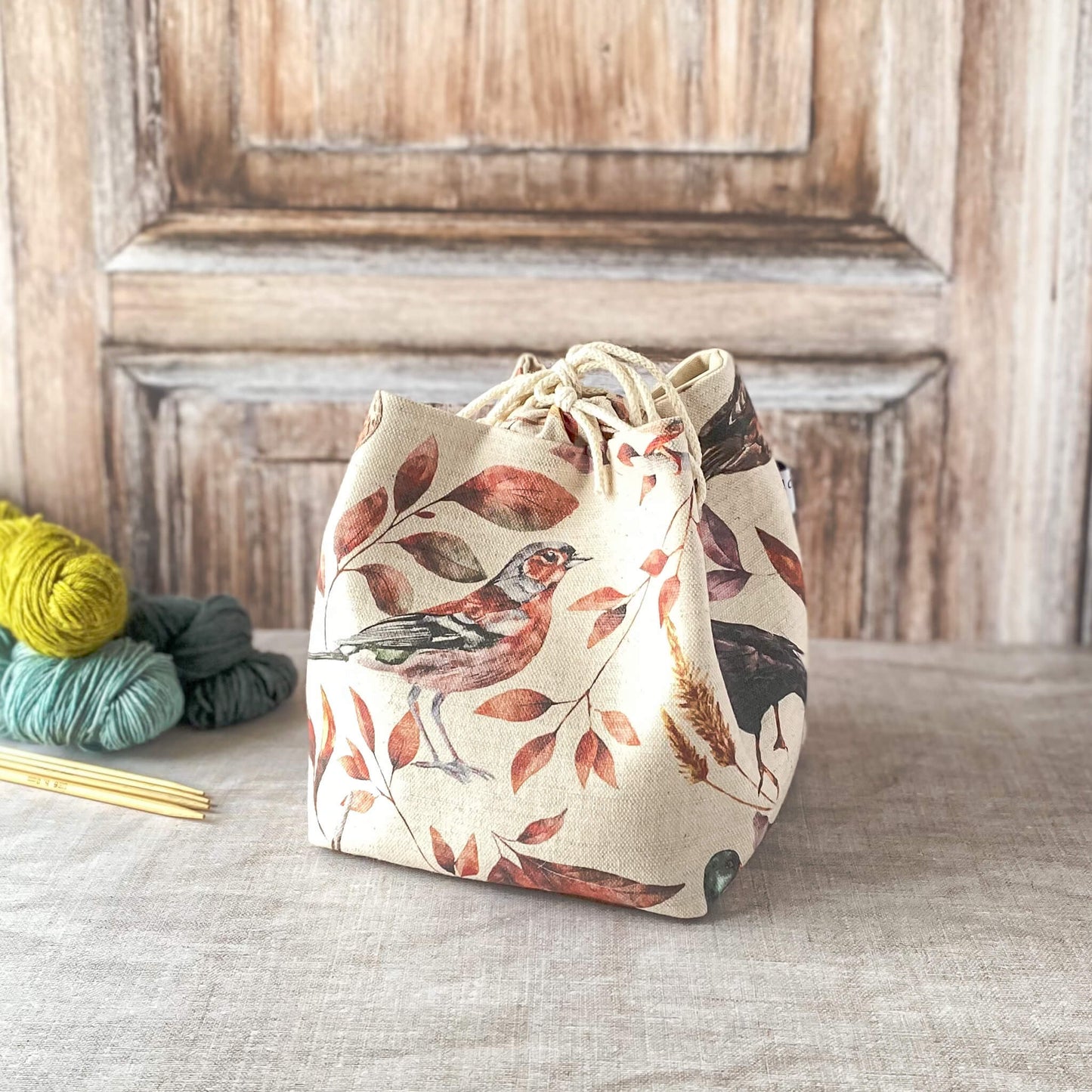 a close-up of a fabric knitting project bag with a bird and leaf design in soft, earthy tones. The bag is secured with a drawstring at the top and placed on a textured surface with visible skeins of yarn and knitting needles nearby. The wooden background adds warmth to the overall aesthetic.