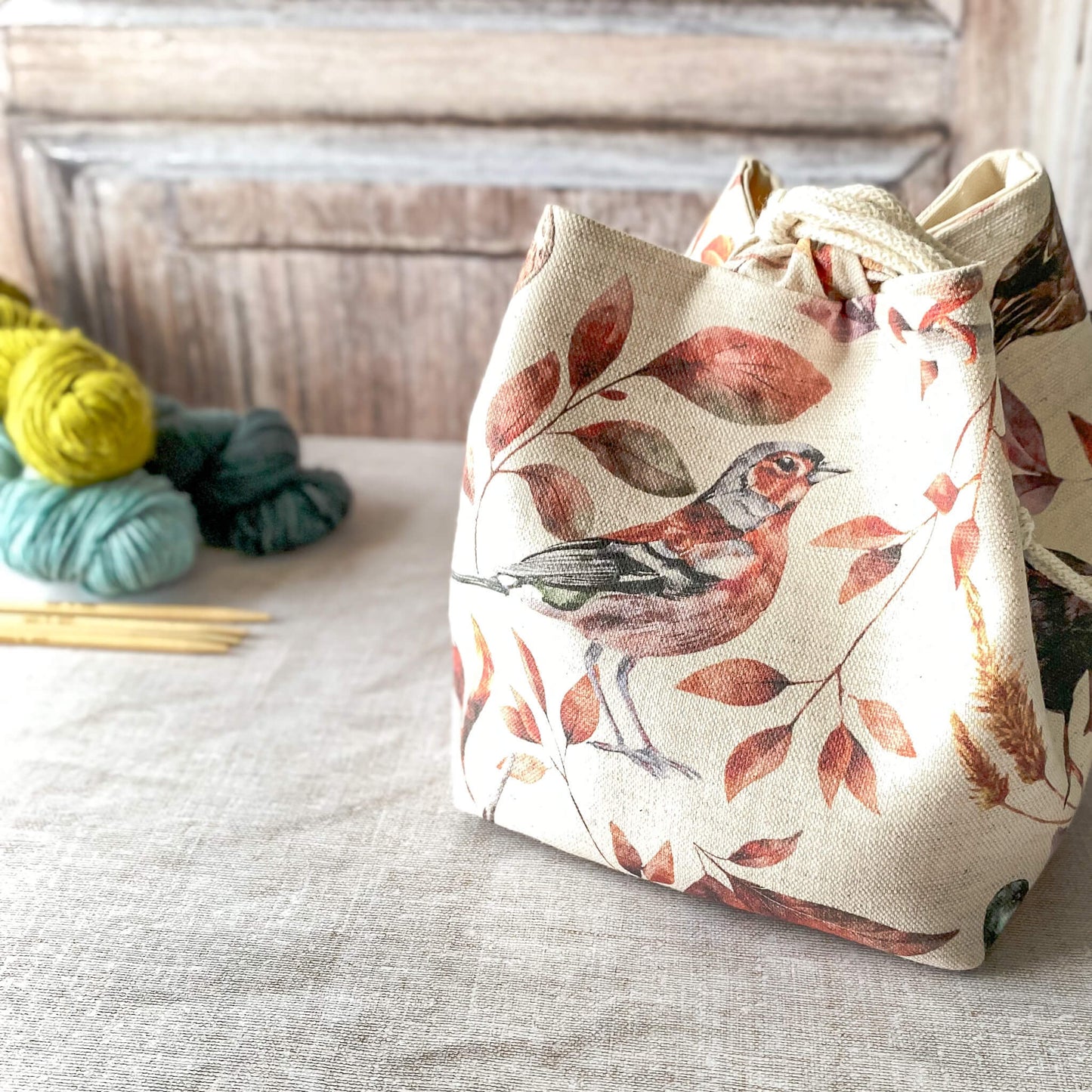 a close-up of a fabric knitting project bag with a bird and leaf print in warm, earthy tones. The bag is cinched at the top with a drawstring, and behind it, you can see a wooden background. Nearby, there are skeins of yarn in shades of green and yellow, along with knitting needles.