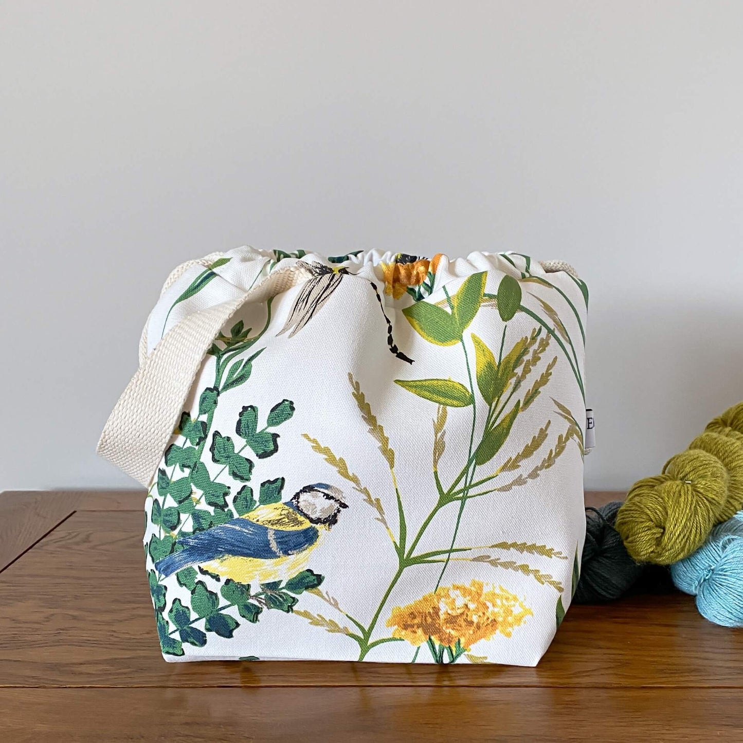 A knitting project bag sits on a table next to three skeins of yarn that are coloured in shades of blue and green. The bag is made froma fabric that shows summer foliage and birds. Prominent is a blue and yellow blue tit.
