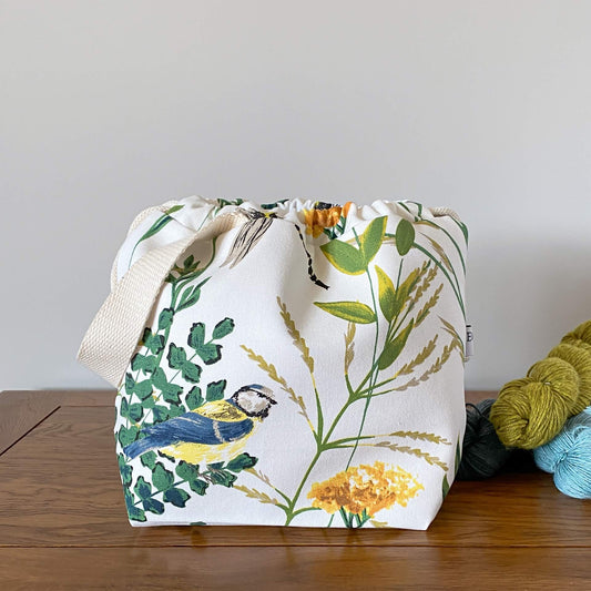 A knitting project bag sits on a table next to three skeins of yarn that are coloured in shades of blue and green. The bag is made froma fabric that shows summer foliage and birds. Prominent is a blue and yellow blue tit. 