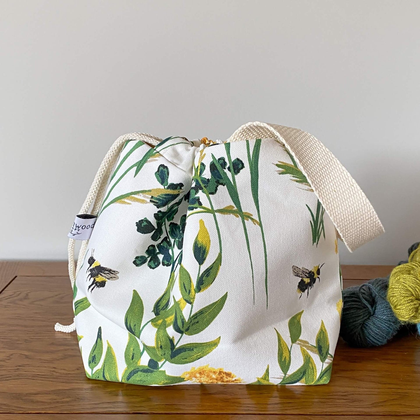 A knitting project bag sits on a table next to three skeins of yarn that are coloured in shades of blue and green. The bag is made froma fabric that shows summer foliage and birds. Prominentare a couple of bumble bees.