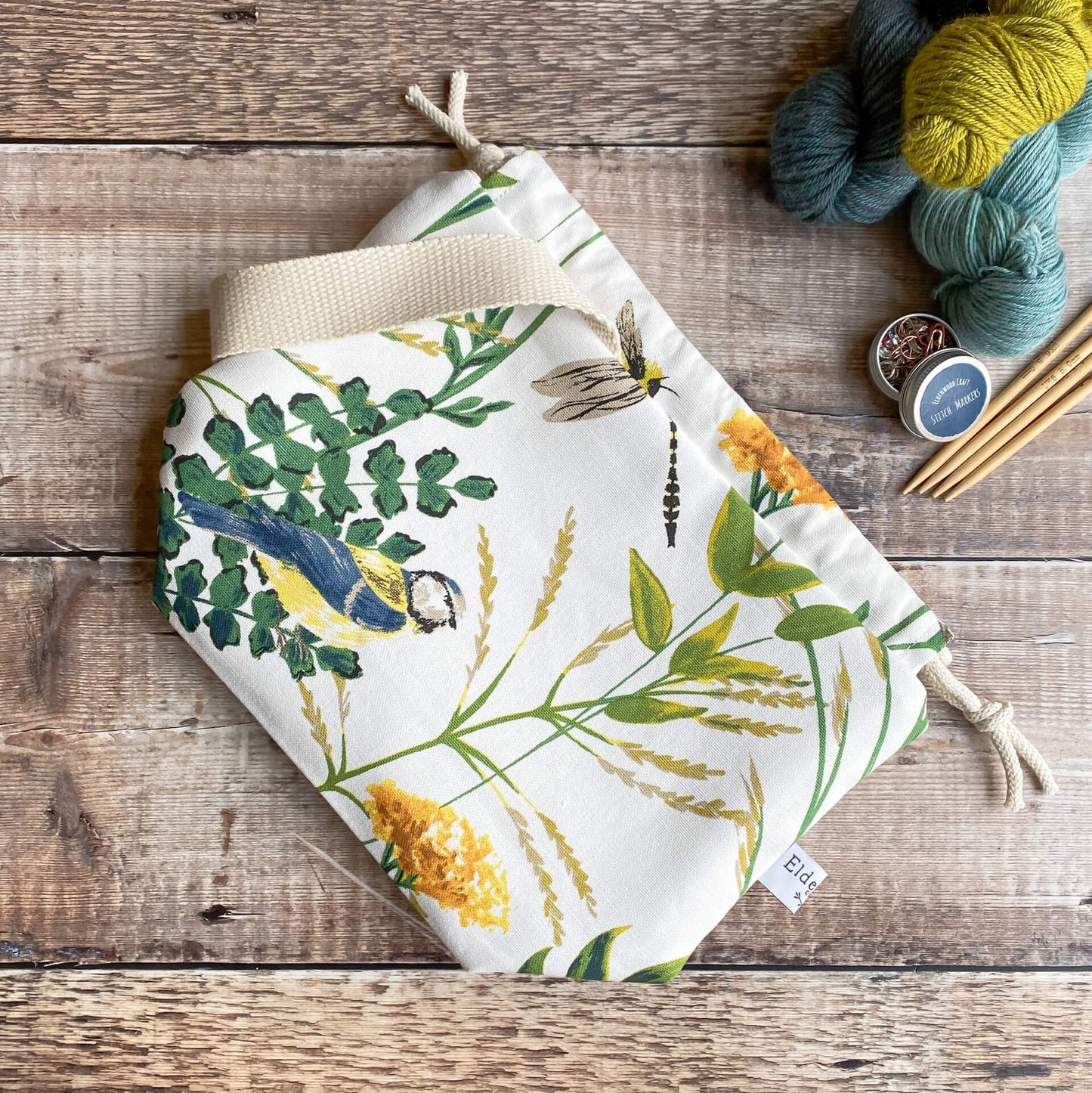 A kntting project bag lies flat on a wooden table top. Next to the bag are three skeins of yarn, some stitch markers and wooden knitting needles.