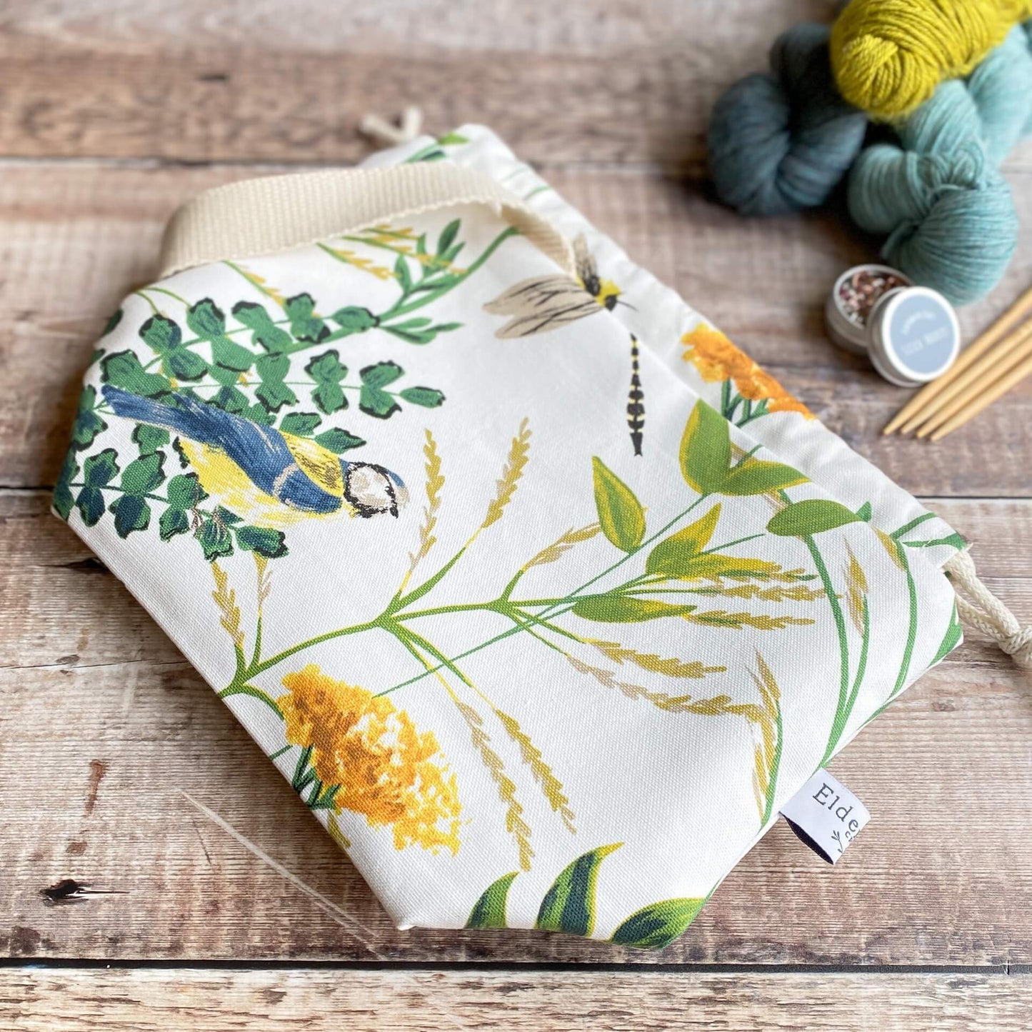 A kntting project bag lies flat on a wooden table top. Next to the bag are three skeins of yarn, some stitch markers and wooden knitting needles. The bag is made from fabric that shows a summery scene with foliage and a blue tit bird prominent.