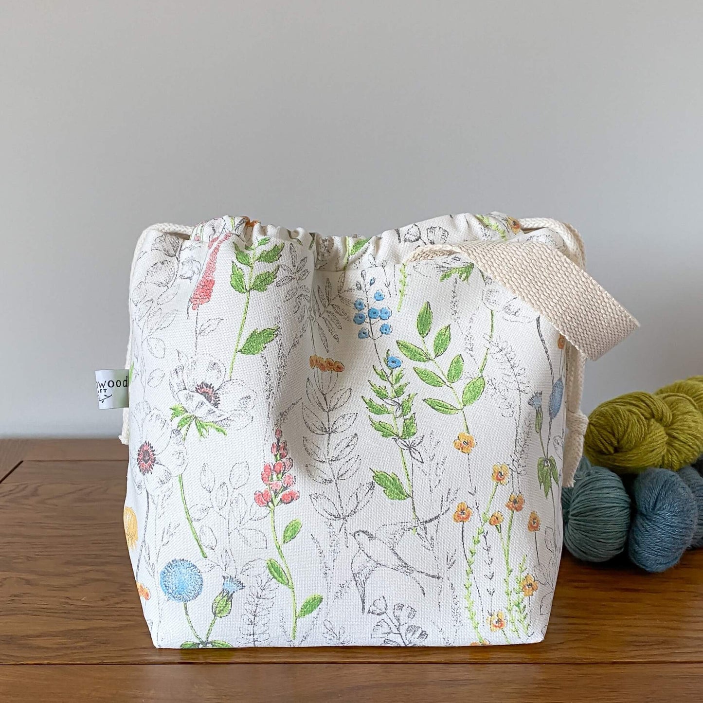 A pretty summery drawstring bag used for knitting and crochet projects sits on a wooden table next to three skeins of yarn. The bag is hand made from a print that shows wild flowers and birds.