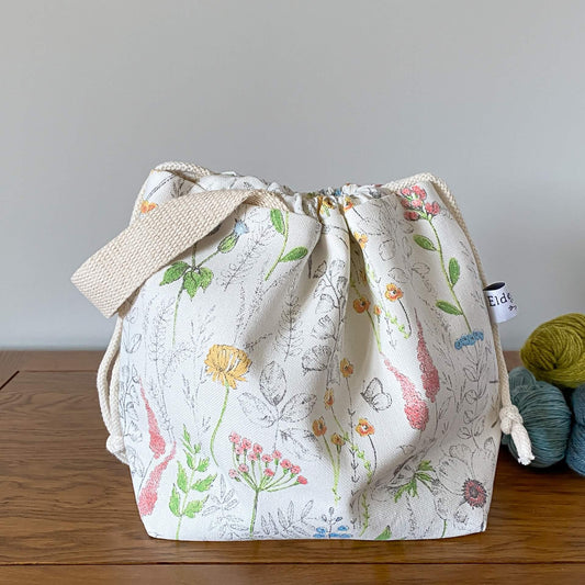 A pretty summery drawstring bag used for knitting and crochet projects sits on a wooden table next to three skeins of yarn. The bag is hand made from a print that shows wild flowers and butterflies. 