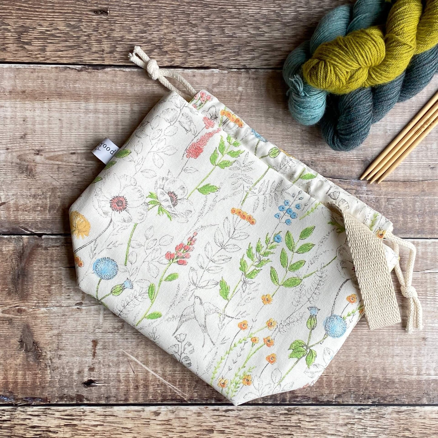 A knitting project bag lies flat on a wooden table next to three skeins of yarn and some wooden knitting needles. The bag is made from a print covered in wildflowers.