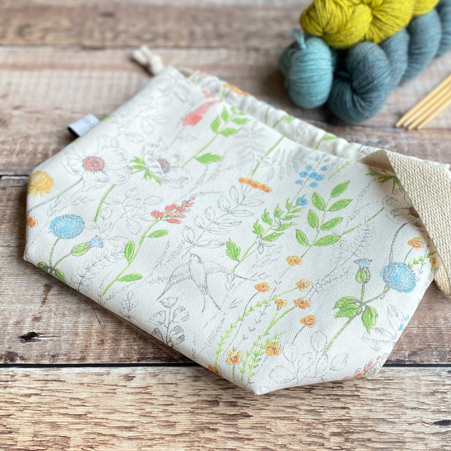 A knitting project bag lies flat on a wooden table with three skeins of yarn and some wooden knitting needles in the background. The bag is made from a print covered in wildflowers and a swallow can also be seen.