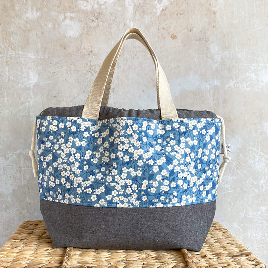A large blue floral project bag for knitting and crochet sits on top of a wicker chest. It is made from Liberty of London fabric and is intended to store large projects like sweaters and blankets.