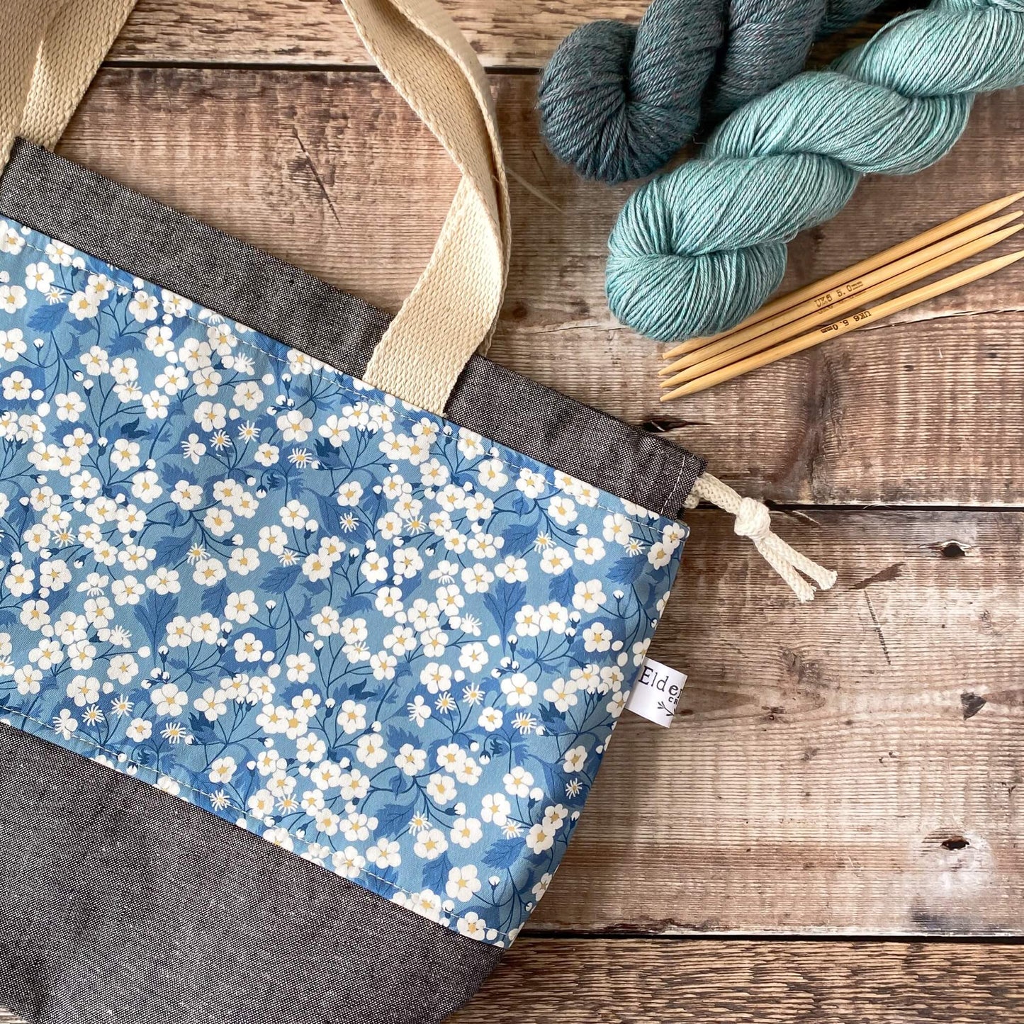 A blue floral project bag for knitting and crochet lies flat on a table. It is a large sized bag designed to store garment and blanket projects.