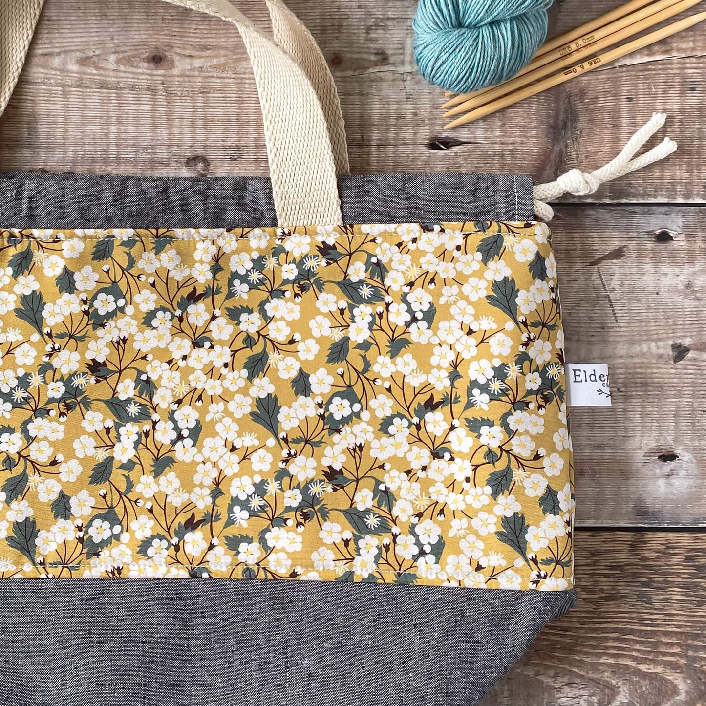 A close up flat lay of a knitting and crochet project bag. The yellow Liberty of London floral fabric is clearly seen as well as the carry handles and the maker's label.