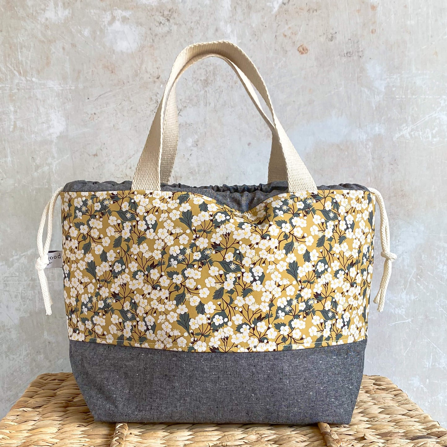 A large project bag for knitting and crochet projects sits on a wicker chest. It is made using yellow Liberty of London floral fabric and has a grey contrasting base and drawstring case. It has two strong carry handles.