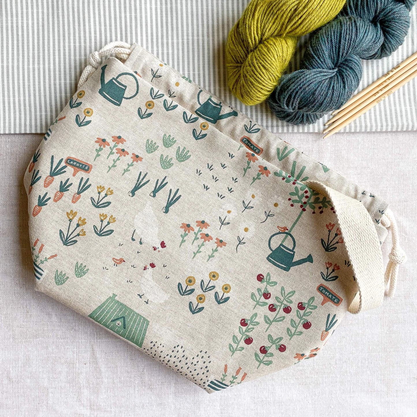 A drawstring bag lies on top of a linen covered table. Next to the bag, which is made out of a whimsical cottage garden fabric, are two skeins of yarn and wooden knitting needles.