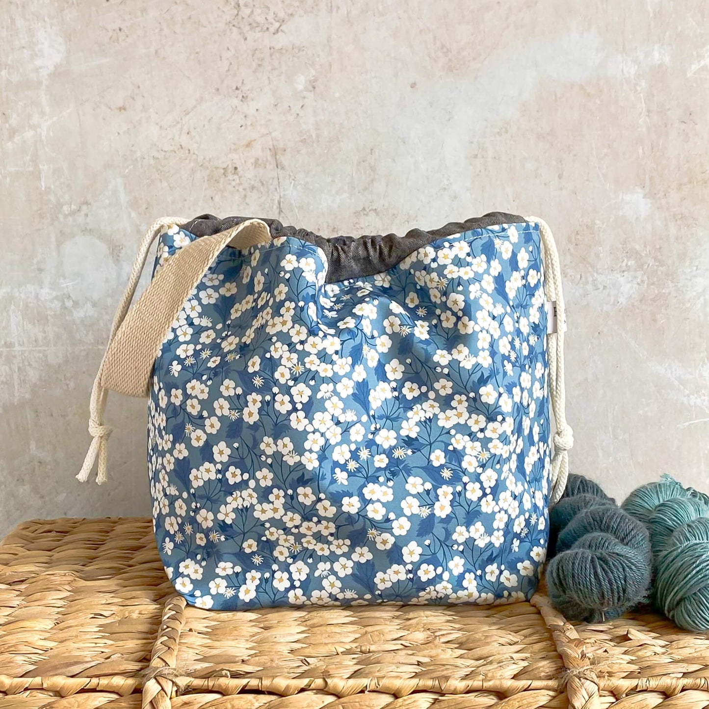 A project bag for knitting and crochet projects sits on a wicker chest. It is made using blue floral Liberty of London fabric. It holds medium sized projects like shawls and has a drawstring closure and a sturdy carry handle.