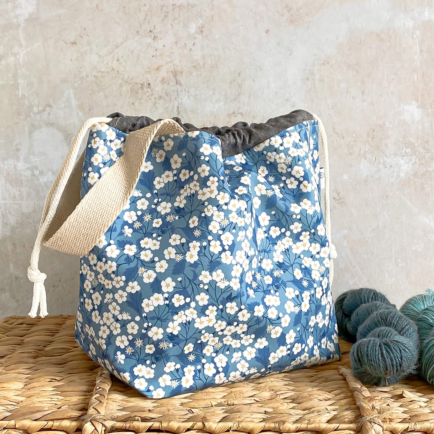 A side view of a blue floral project bag designed to hold knitting and crochet projects like shawls and light garments. It's carry handle is clearly visible as well as the drawstrings.