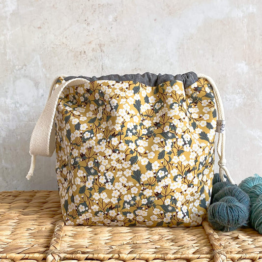 A medium sized knitting and crochet project bag sits on a wicker chest. It is made using yellow floral fabric from Liberty of London. It will hold projects like shawls and small garments.