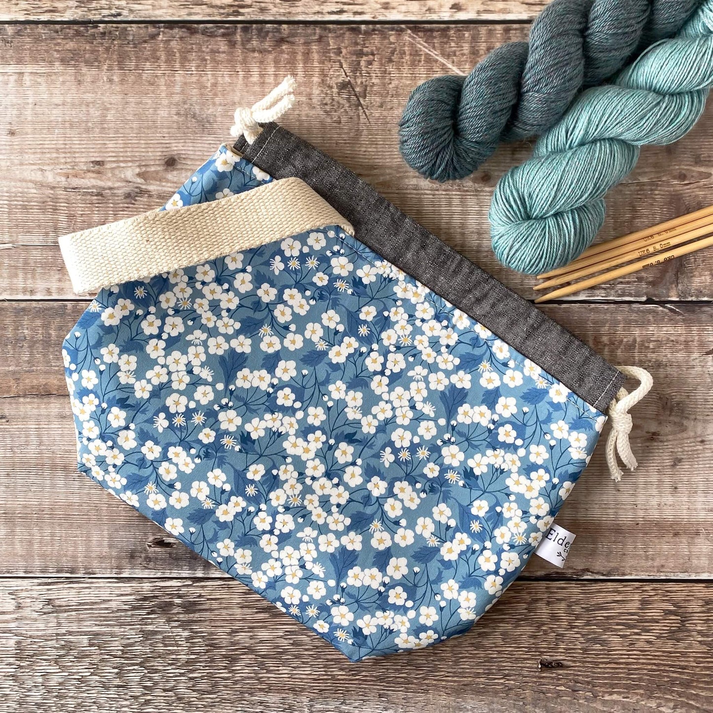A flat lay of a blue floral project bag for knitters and crocheters. It has a handle and closes by a drawstring.