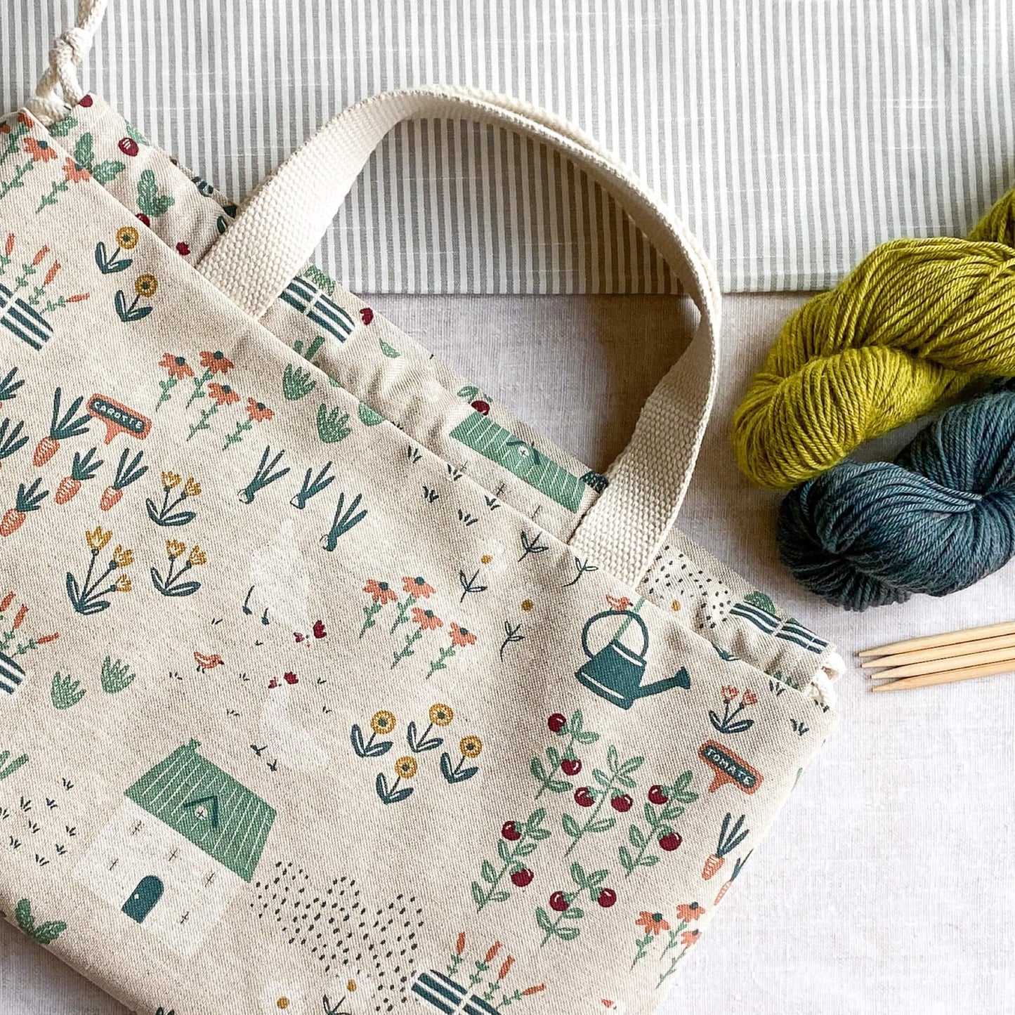 A large drawstring project bag for knitting lies flat on a linen covered table. Next to the bag are two skeins of yarn. One is blue and the other green. Next to the yarn lies four wooden knitting needles.