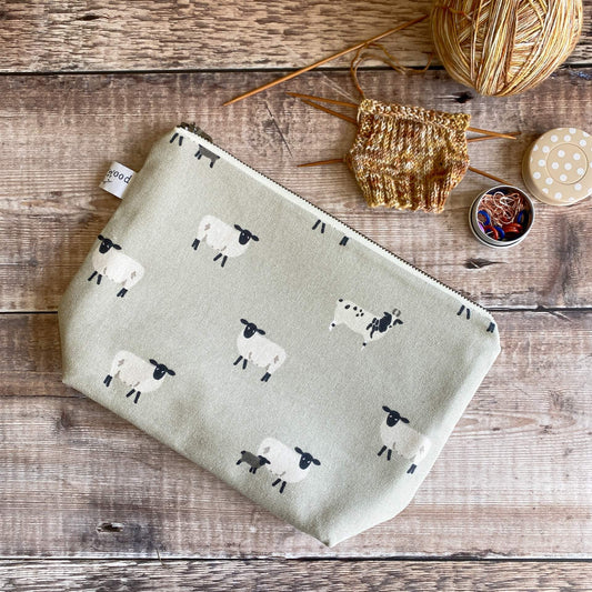 A small zipped pouch lies on a wooden table next to some knitting. The bag is made from a sheep print fabric on a grey background. 