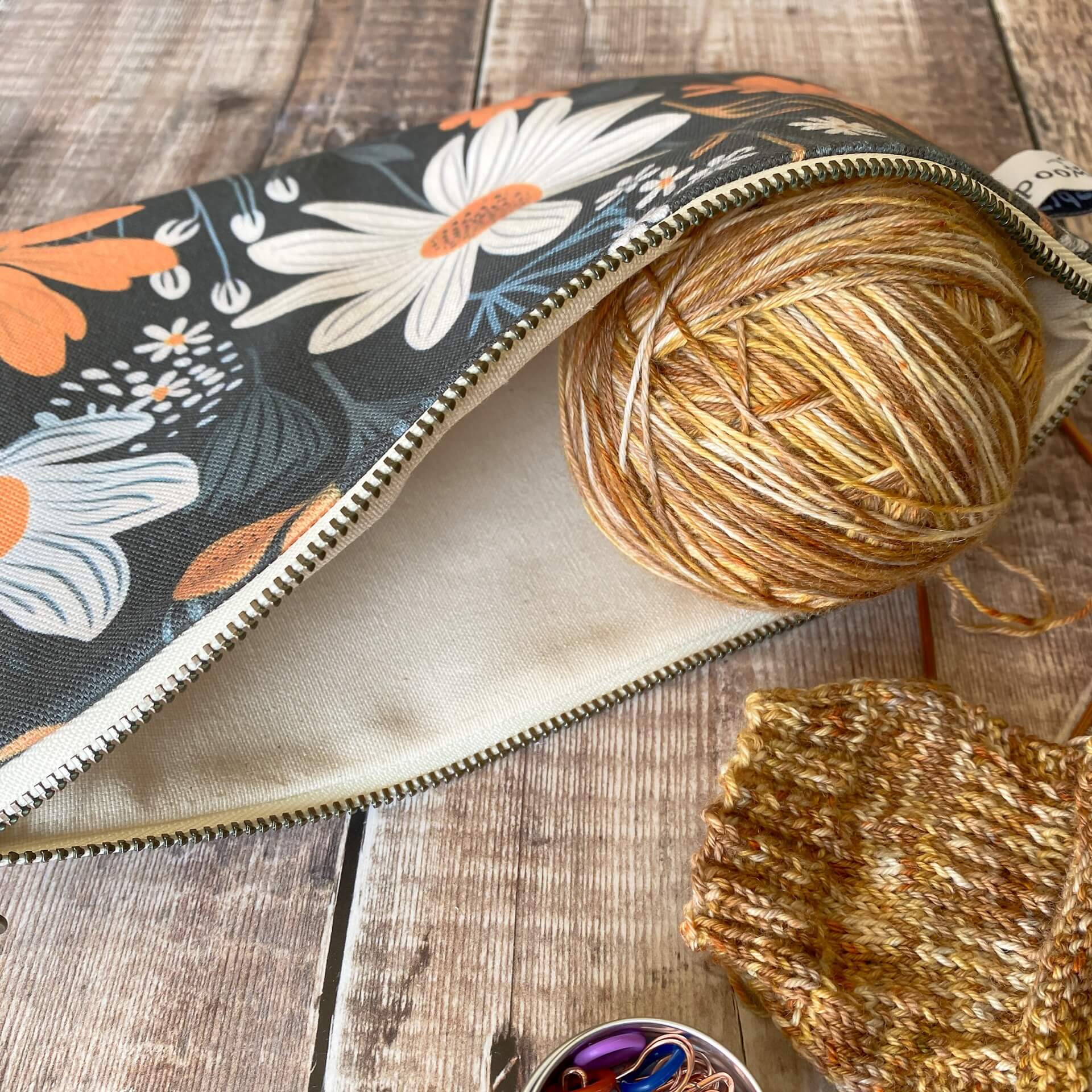 An opened zipped pouch lies on a wooden table. In the opening sits a  ball of brown toned yarns next to a knitting project in progress. 