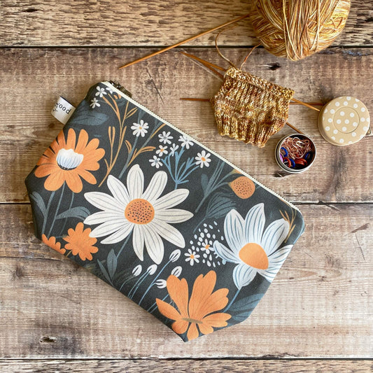 A small zipped pouch featuring a bold and dark floral print lies on a wooden table next to some knitting. 