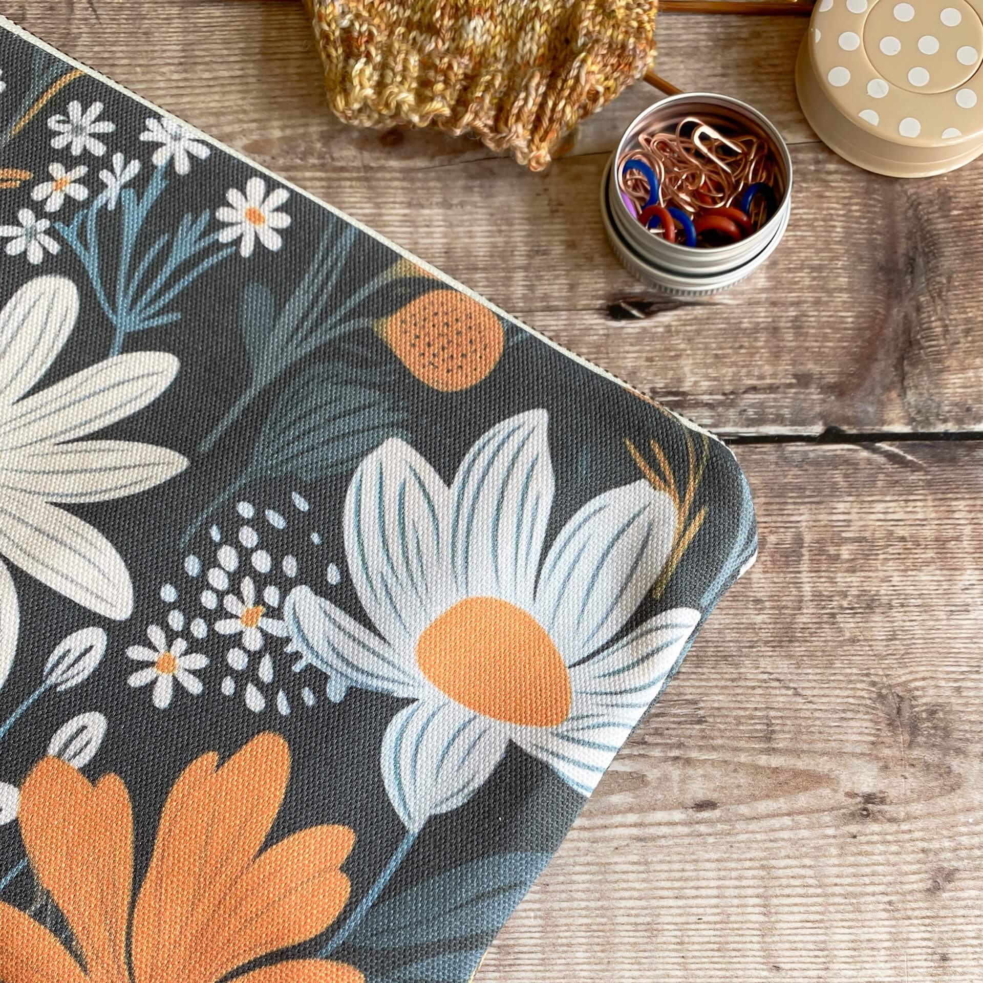 A close up of a small zipped pouch featuring a bold and dark floral print lies on a wooden table next to some knitting. 