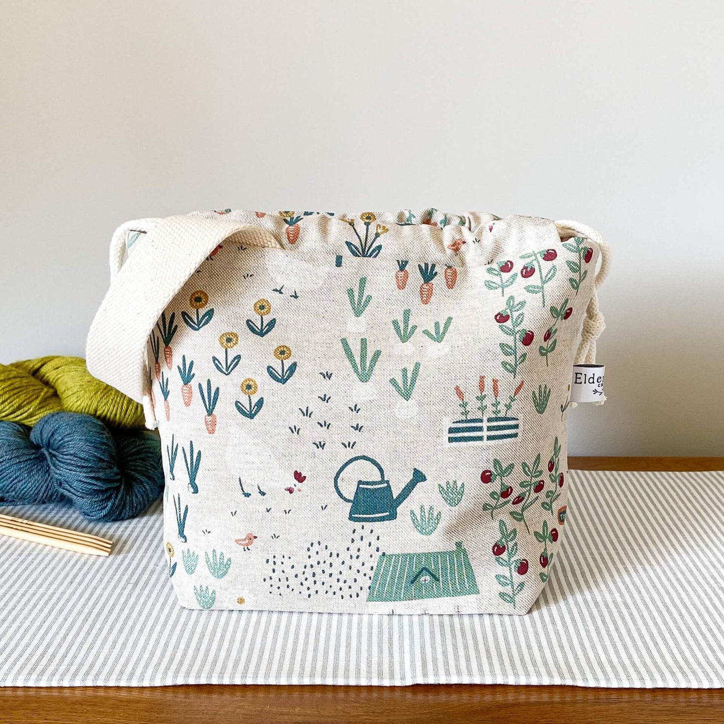 A knitting project bag sits on a table top. Behind the bag are some skeins of yarn in different colours neatly arranged. Also in view are some wooden knitting needles. The bag is at the centre of the image and is made from a fabric that depicts a cottage garden.