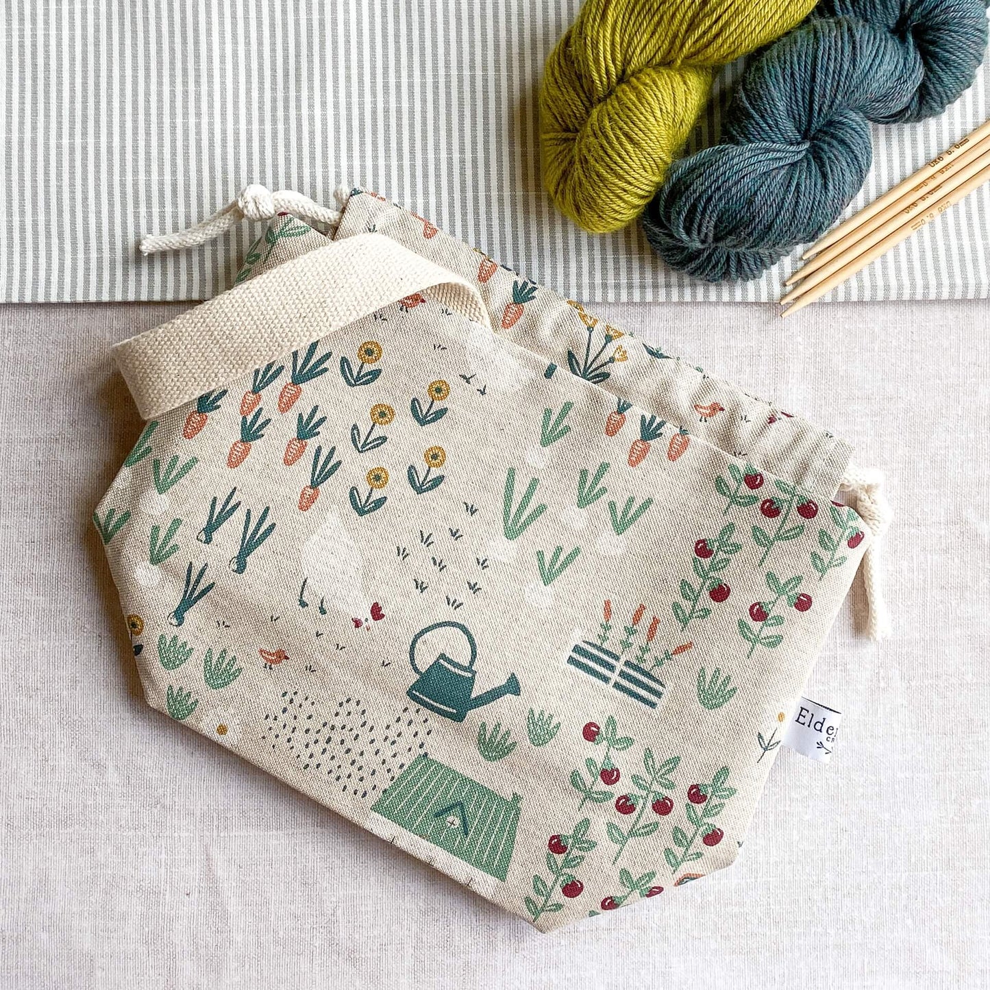 A bag showing images of chickens and flowers and vegetables in a whimsical style is lying on a table top. Just in shot are two skeins of yarn, one green and one blue, alongside four wooden knitting needles.