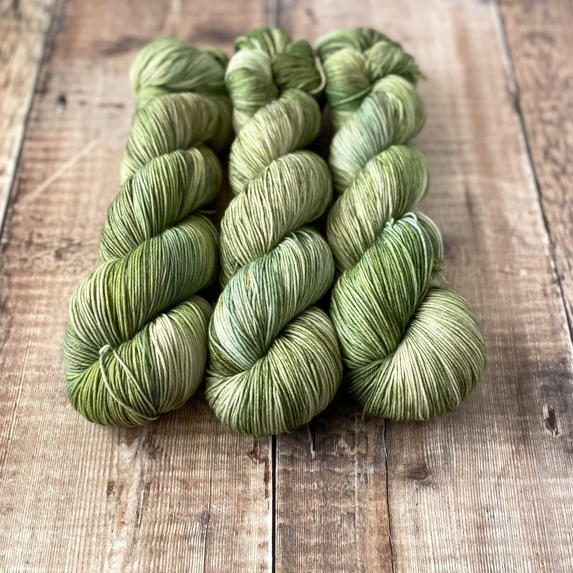 3 skeins of variegated yarn in greens and blues and greys, sitting in a line on a wooden surface