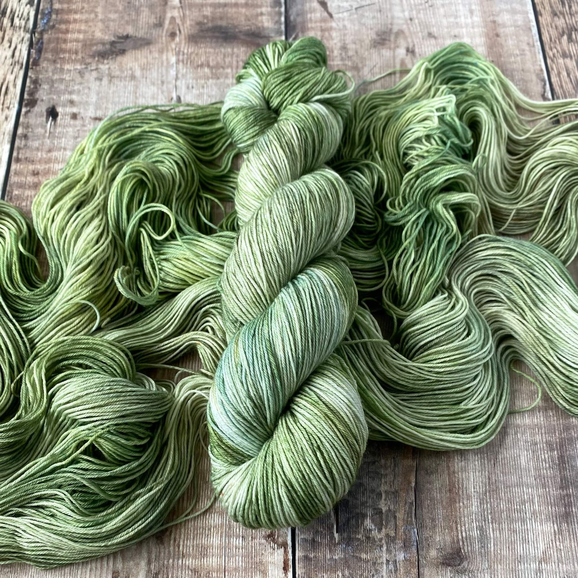 A skein of hand dyed yarn in green and grey, sitting on a skein of the same yarn unwound on a wooden surface