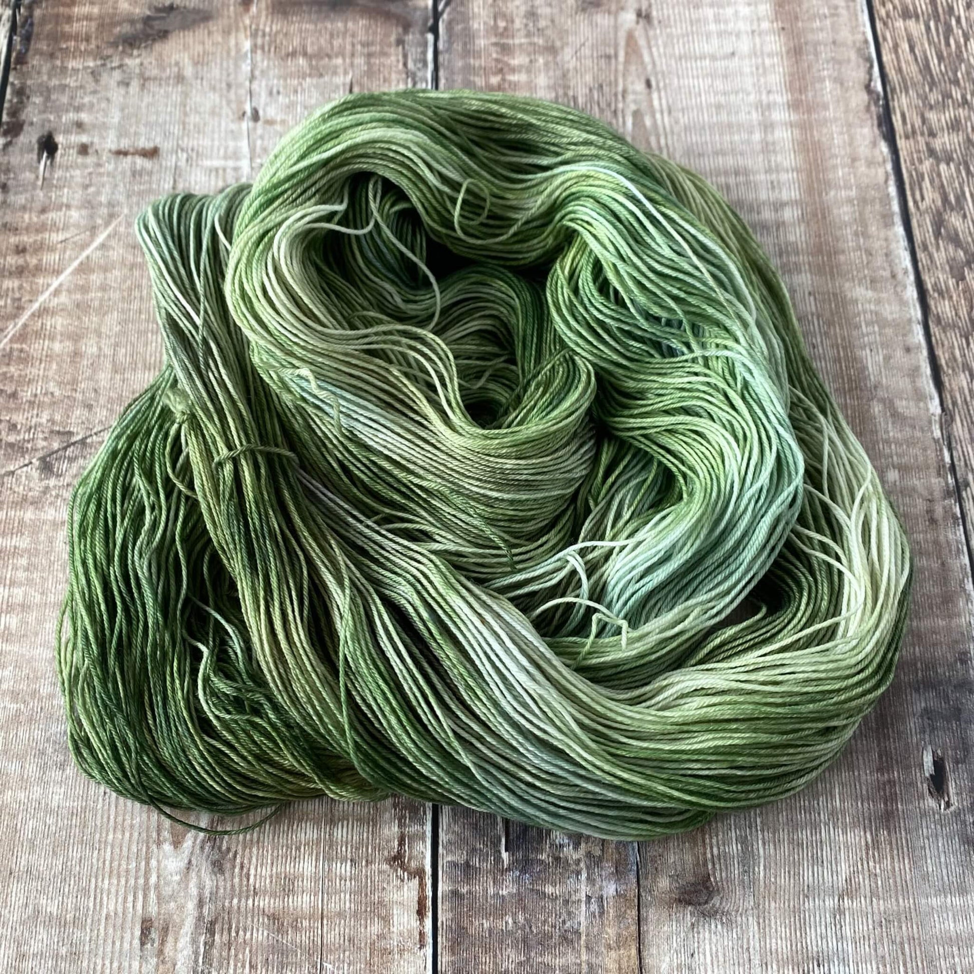 A skein of green and grey hand dyed yarn coiled in a circle on a wooden surface