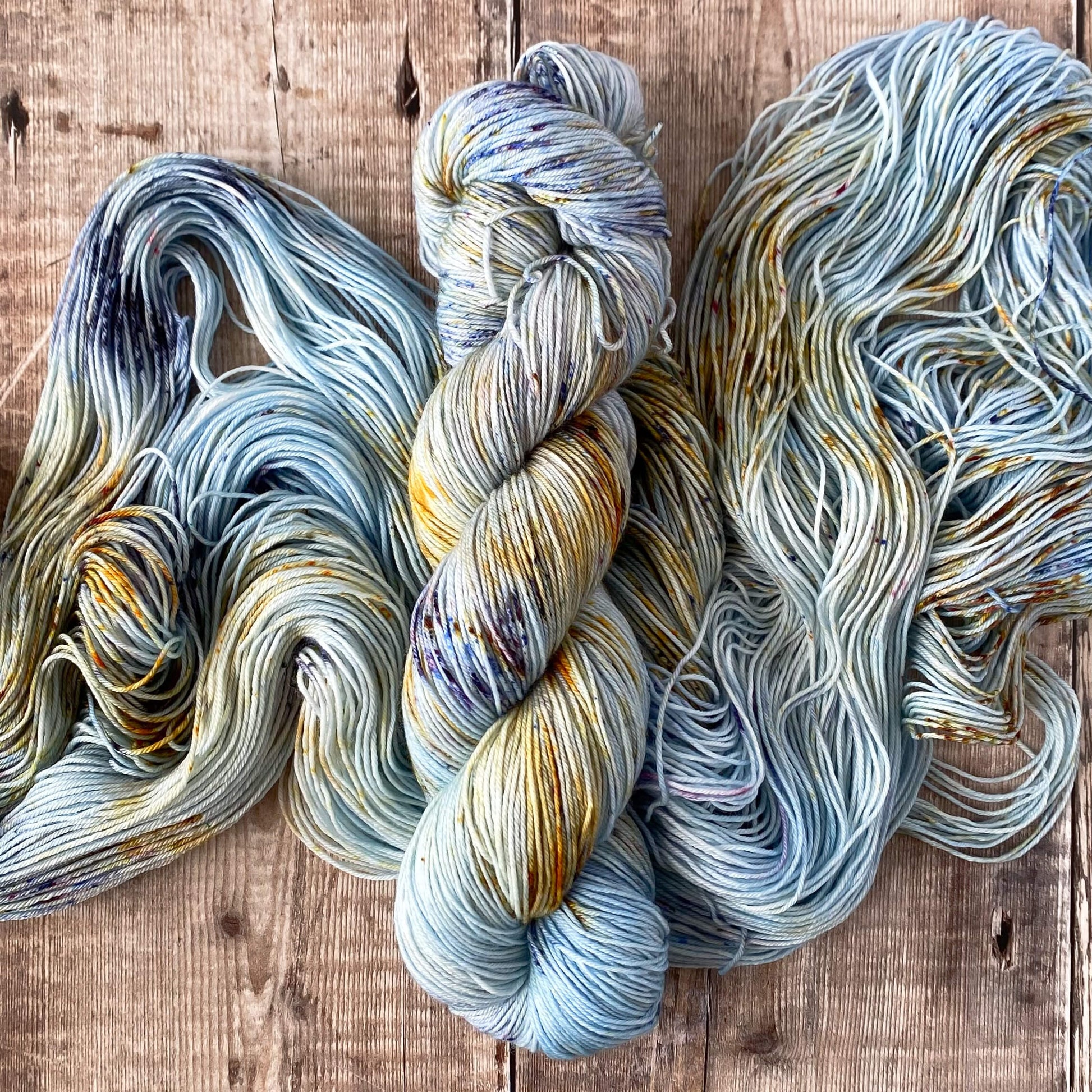 A skein of variegated yarn in blue, orange, brown, sitting on an unwound skein in the same colours, both on a wooden surface
