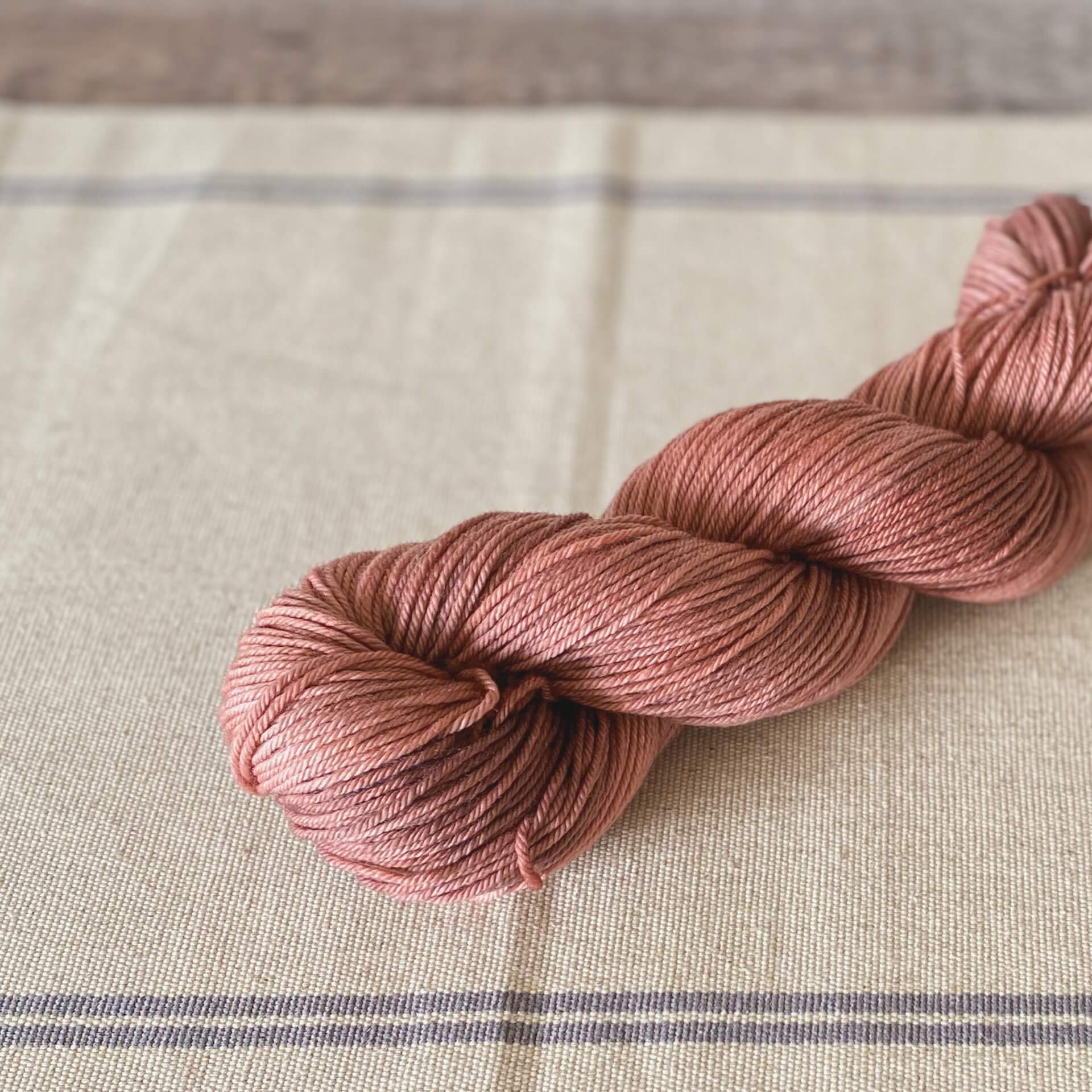 A wound up skein of yarn sits on a linen table runner. The yarn is coloured a deep pink. 