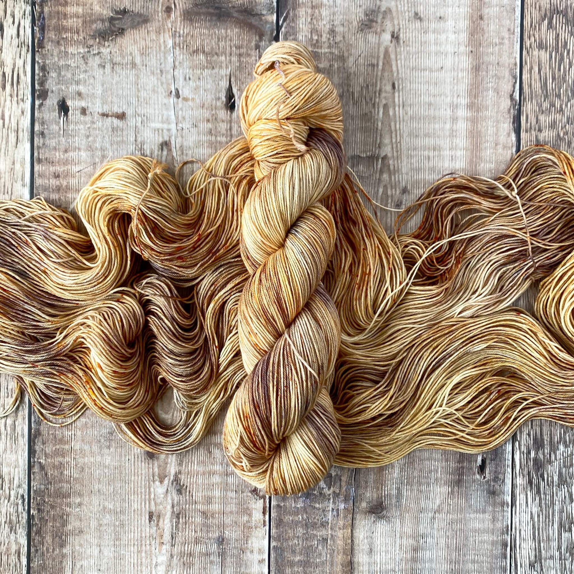 A variegated skein of hand dyed yarn coloured in browns and greys sits on a skein of the same yarn, unwound on a wooden surface.