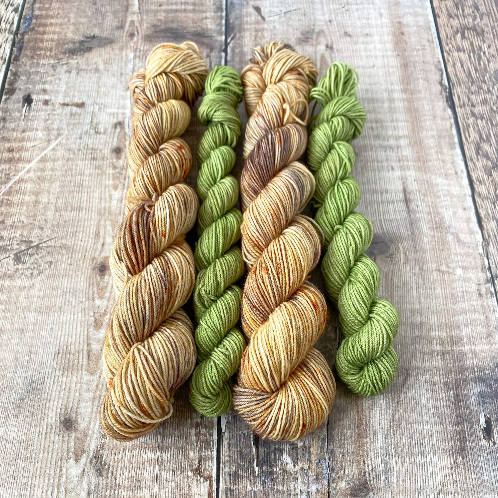 Two hand dyed sock sets sit in a row on a wooden surface. Two larger skeins are coloured brown and grey and two smaller skeins are coloured green.
