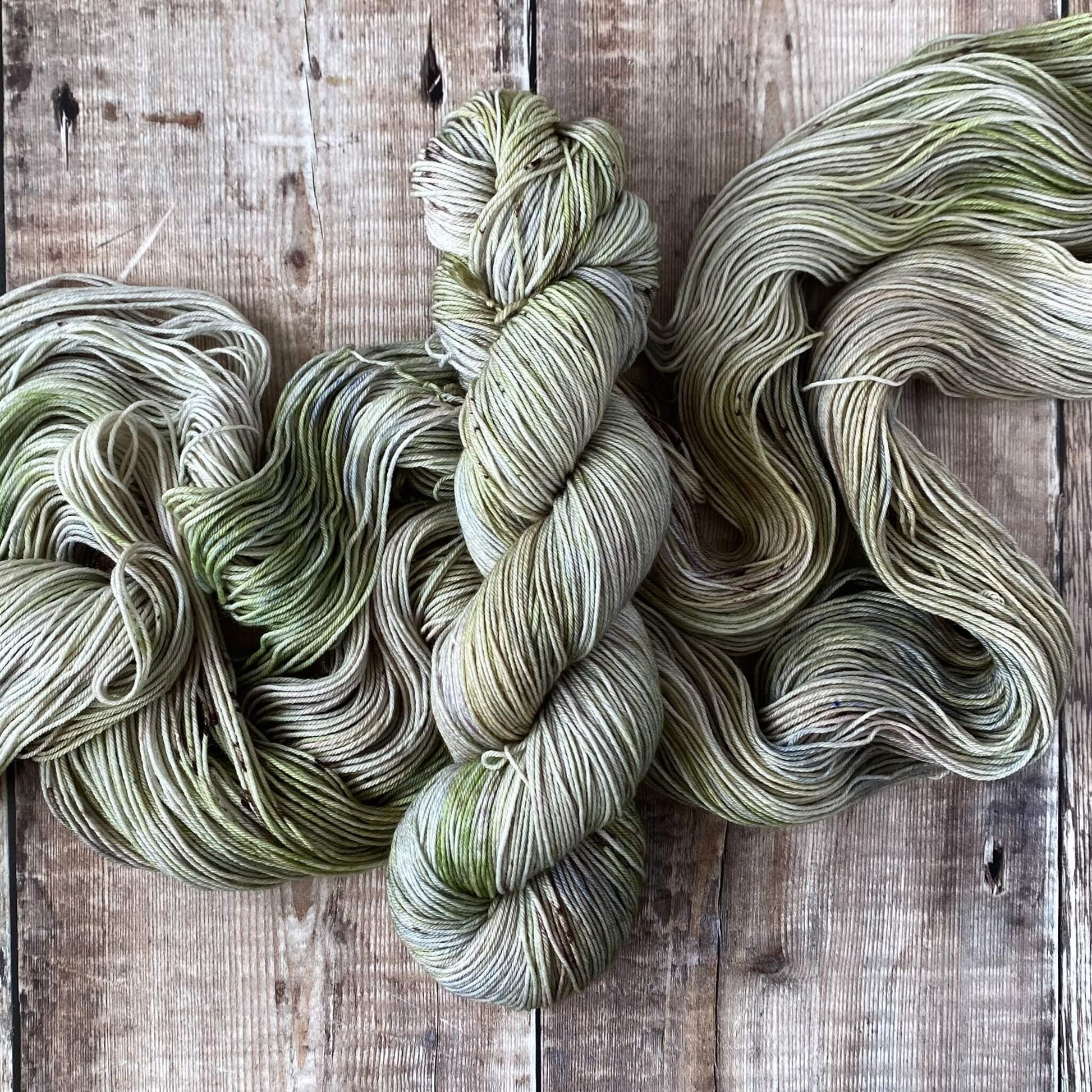 A skein of hand dyed yarn in green and grey wound into a hank a lying on an unwound skein.