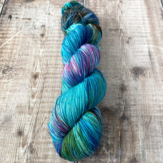 A skein of hand dyed yarn in blues, greens and pinks seen on a wooden surface.