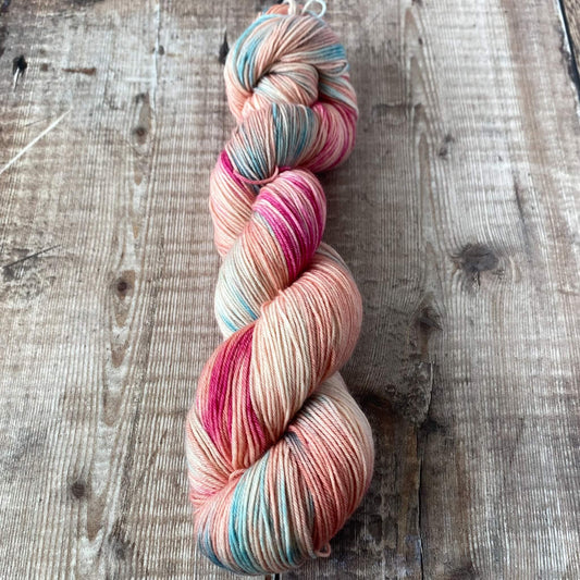 A variegated skein of hand dyed yarn seen on a wooden surface. The skein is coloured with pinks and blues.