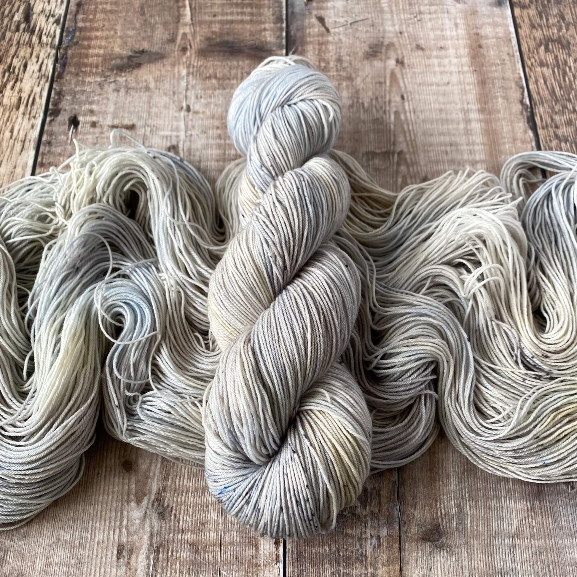 A skein of hand dyed yarn wound in a hank lies on top of an unwound skein. Both skeins are pastel blues and greys.