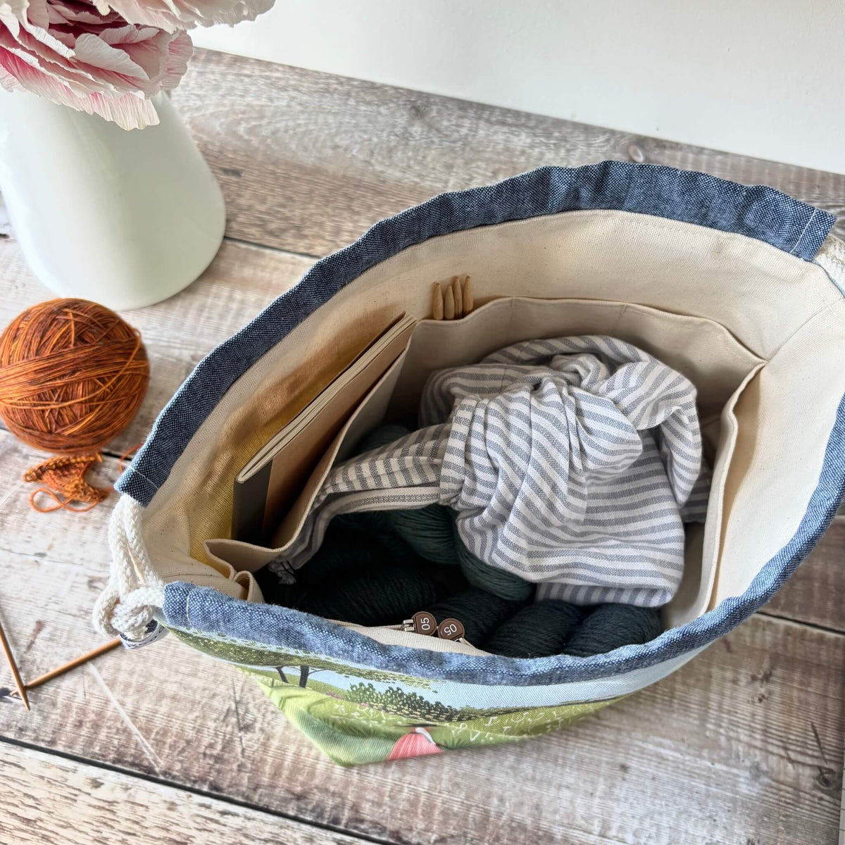 A fabric storage basket with a cream-colored interior, shown filled with knitting supplies on a rustic wooden table. A white vase with pink flowers and a ball of orange yarn are visible in the background.