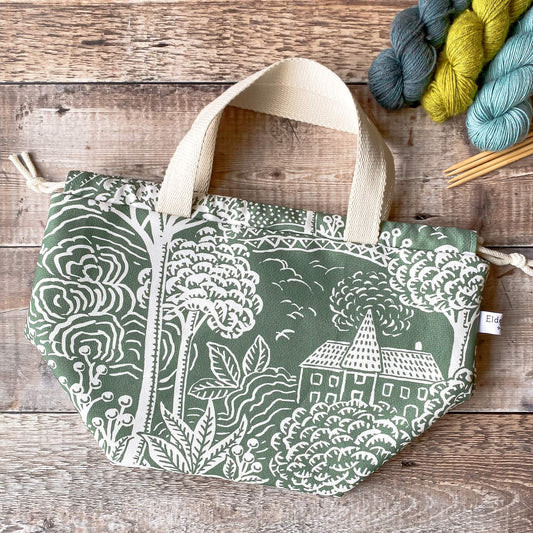 A large sized green drawstring project bag with a print of a house and gardens lies on a wooden surface next to three skeins of yarn and wooden knitting needles.