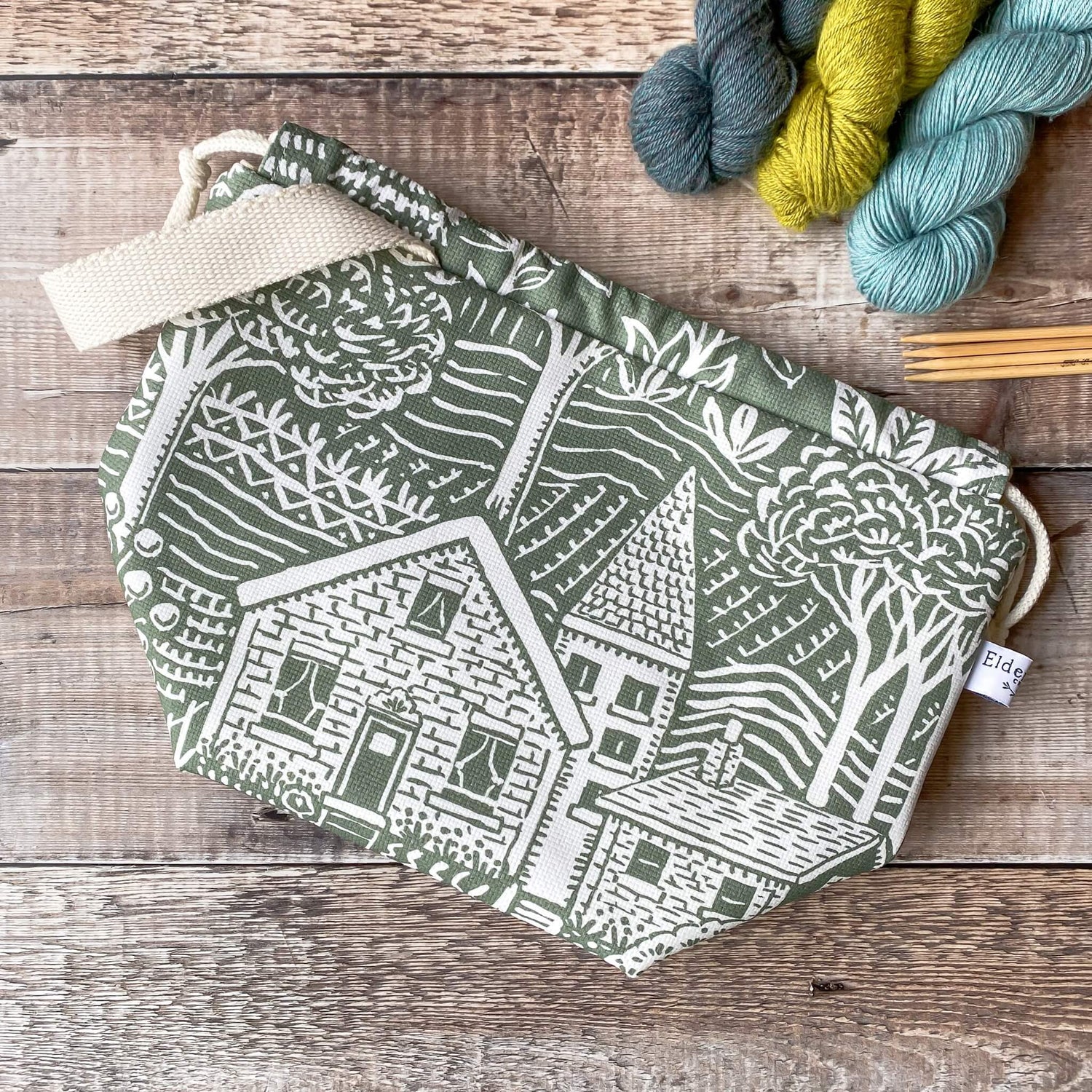A medium sized green drawstring project bag with a print of a house and gardens lies on a wooden surface next to three skeins of yarn and wooden knitting needles.