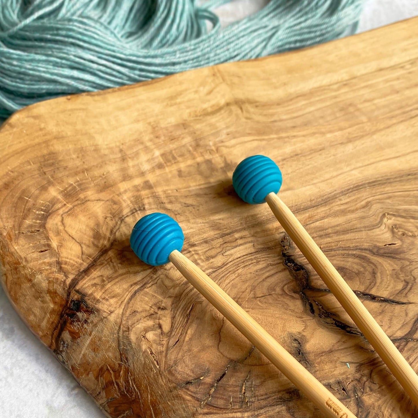 Two blue silicone balls sit attached to the end of two knitting needles. The needles lie on a wooden surface next to some pale green yarn.