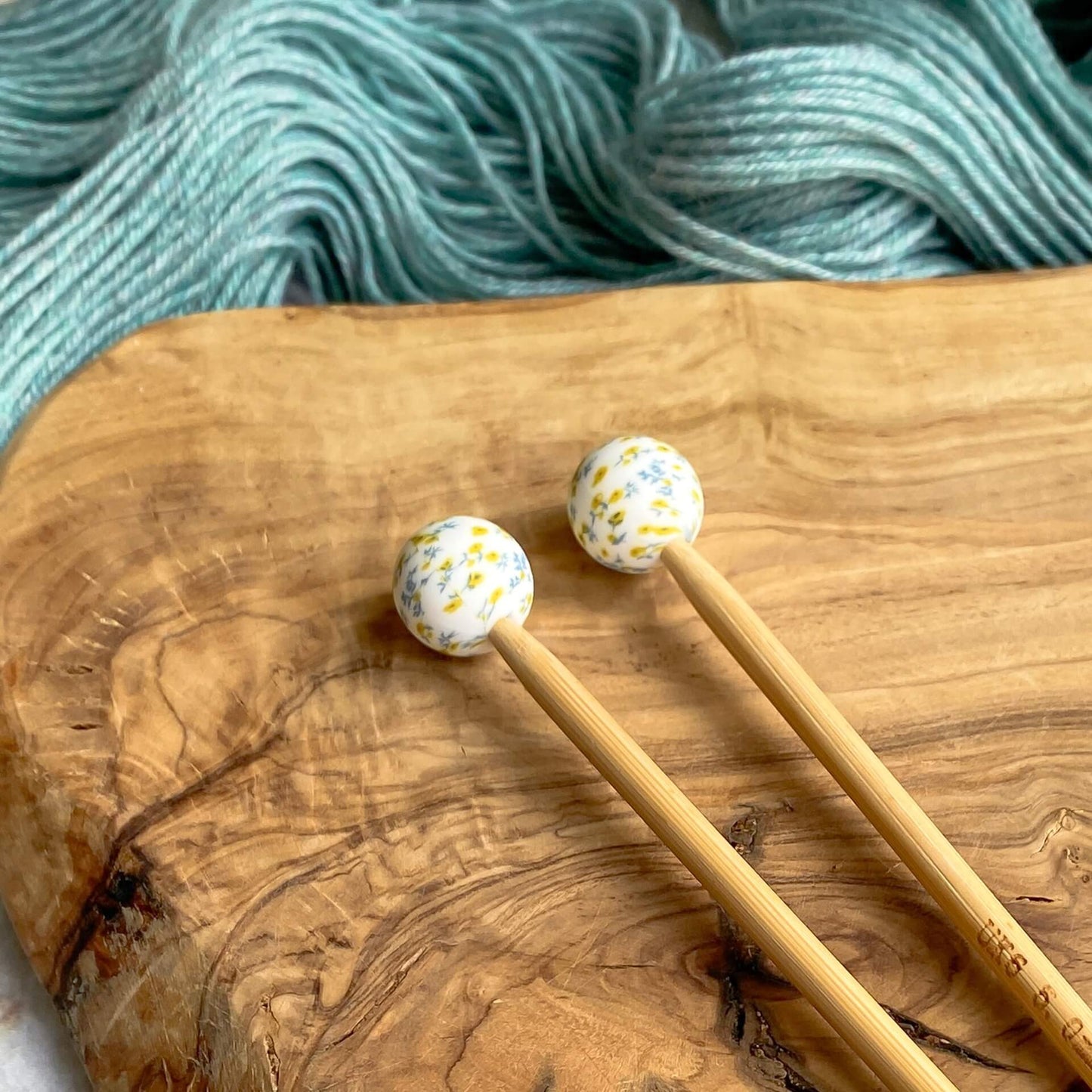 Two silicone balls printed with a ditsy floral pattern sit attached to the end of two knitting needles. The needles lie on a wooden surface next to some pale green yarn.