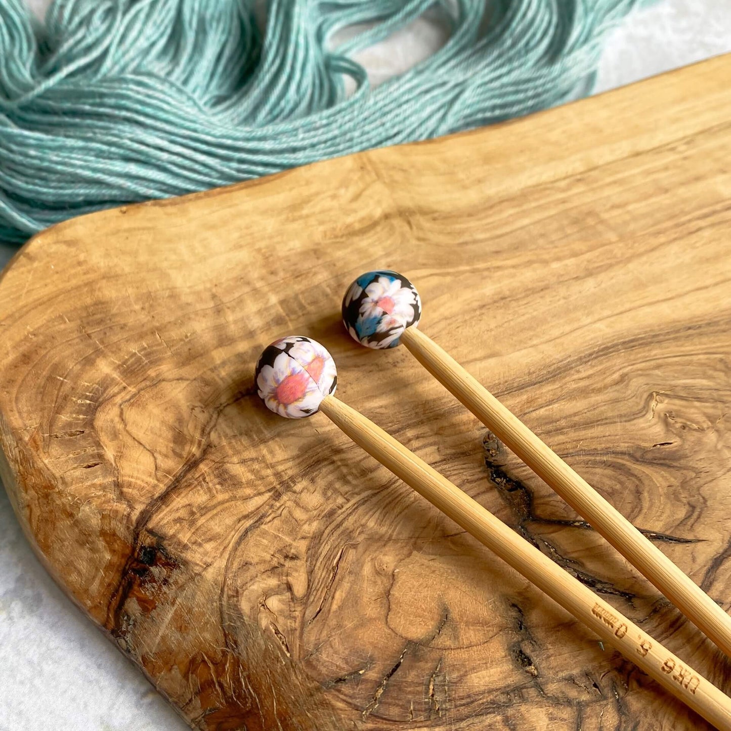 Two silicone balls printed with a bold floral pattern sit attached to the end of two knitting needles. The needles lie on a wooden surface next to some pale green yarn.
