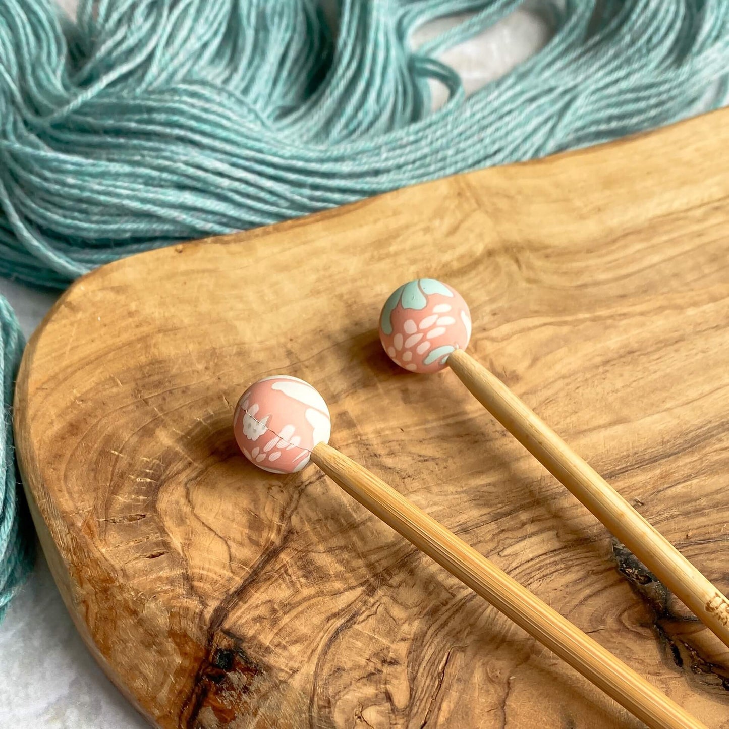 Two silicone balls printed with a calm pink floral pattern sit attached to the end of two knitting needles. The needles lie on a wooden surface next to some pale green yarn.