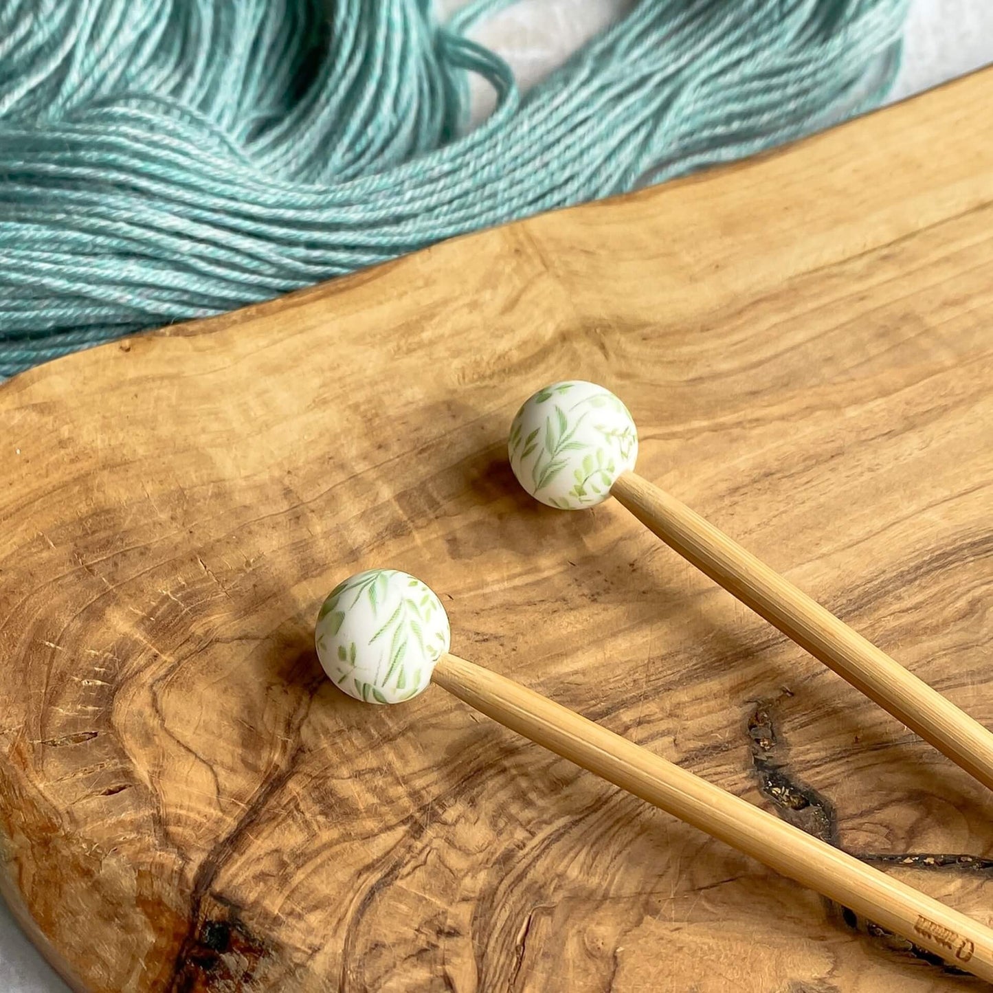 Two silicone balls printed with a green leafy pattern sit attached to the end of two knitting needles. The needles lie on a wooden surface next to some pale green yarn.